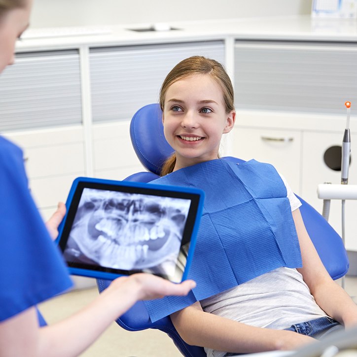 Smiling young girl in dental chair while dentist reviews digital x-rays